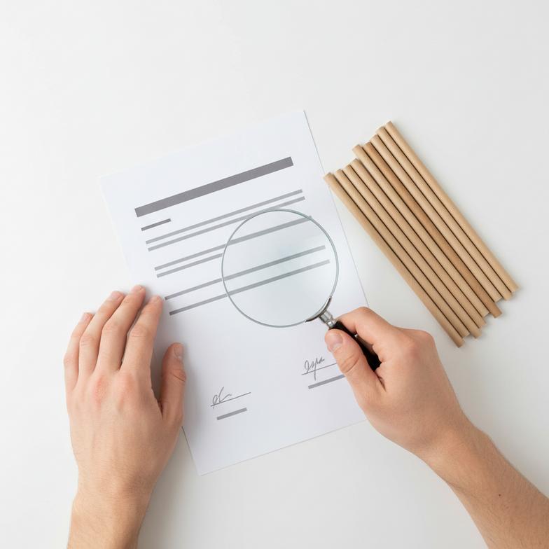 An image of a person with a magnifying glass closely examining a compliance document.